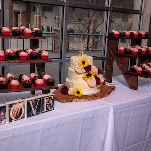 May include: A three-tiered wedding cake with white frosting and sunflowers, surrounded by cupcakes with pink and brown frosting. The cake is on a wooden slice, and the cupcakes are on wooden shelves. The table is covered in a white tablecloth, and there are wooden blocks spelling "LOVE" in front of the cake.