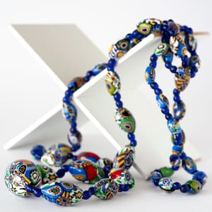 Op de afbeelding: Een kleurrijke ketting met grote, kleurrijke kralen in blauw, groen, rood en geel. De ketting is geregen op een donkerblauw koord. De kralen hebben ingewikkelde ontwerpen en worden gepresenteerd tegen een witte achtergrond.