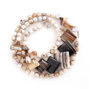 May include: A necklace with a mix of round and rectangular beads in shades of brown, cream, and black. The necklace has a silver clasp and is displayed on a white background. The beads have a polished finish.