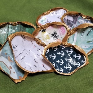 May include: A collection of eight oyster shells with gold trim. Each shell is decorated with a different patterned paper, including a map, pineapples, tropical leaves, and anchors.