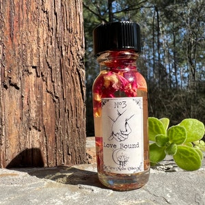 May include: A small glass bottle filled with a clear liquid and rose petals. The bottle has a black label with the text "No. 3 Love Bound" and the words "New Moon Vibes".