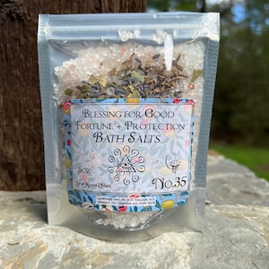 May include: A clear plastic pouch filled with white bath salts, herbs, and flowers. The pouch has a label that reads "Blessing for Good Fortune + Protection Bath Salts" with a design of an eye inside a triangle and a crescent moon with a lotus flower. The label also says "5oz New Moon Vibes No.35".