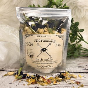 May include: A small clear plastic bag filled with bath salts and dried herbs. The bag has a label that reads "New Moon Vibes" and "Uncrossing No. 14" with a black and white illustration of an eye and crossed arrows. The label also states "Bath salts" and lists the ingredients: Dead Sea Salt, Himalayan Salt, Essential Oil, Fragrance and Dried Herbs. The bag is 5 oz.