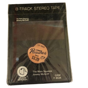 May include: A black 8-track stereo tape cassette with a brown and orange image on the front. The cassette is shrink-wrapped and has a price tag that says "Peaches List Price £3.29". The title of the album is "The Main Squeeze" by "Jimmy McGriff" and the label is "GRM F 8534".