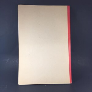 May include: A blank, off-white book with a red spine. The book is closed and lying flat on a dark gray surface.