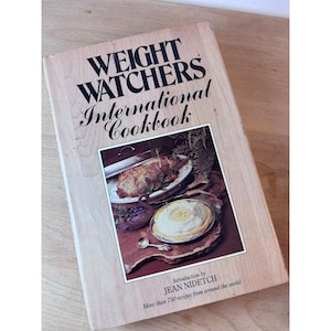 Peut inclure: Un ancien "Weight Watchers International Cookbook" avec une couverture en bois clair. Le titre est en lettres noires en gras. La couverture présente une photo d'un repas. Le livre comprend plus de 750 recettes du monde entier.