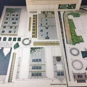 May include: A paper model kit of Neuschwanstein Castle, a famous German castle. The kit includes detailed instructions and pieces to build the castle, including the towers, walls, and windows. The castle is depicted in a light gray color with green accents.