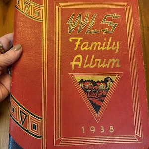 Könnte beinhalten: Rotes, ledergebundenes Familienalbum mit goldenen Akzenten und dem Text "WLS Family Album 1938". Der Umschlag zeigt eine Schwarzweißillustration einer Farmszene mit einem Haus, einem Stall und einer Person, die ein Feld pflügt.