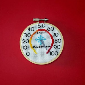 May include: A white circular cross-stitch with a red, orange, yellow, and blue color gradient. The cross-stitch is a thermometer with the words "spirit" and "clousometer" and a snowflake design. The thermometer needle points to 50 degrees.