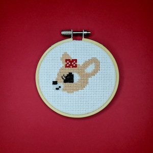 May include: A white embroidery hoop with a cross-stitch design of a fawn with a red bow. The fawn is looking to the left and has a black eye.