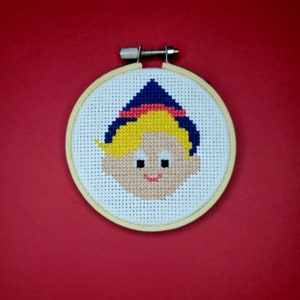 Op de afbeelding: Een borduurring met een kruissteek met een cartoon-elf ontwerp. De elf heeft blond haar, een blauwe hoed met een rode band en een glimlachend gezicht.