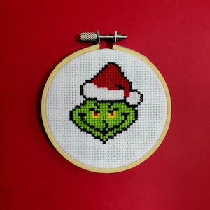 May include: A green Grinch face with a red Santa hat embroidered on white fabric in a wooden embroidery hoop.