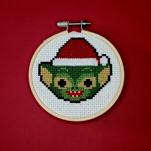 May include: A green and red cross-stitch embroidery of a creature wearing a Santa hat, framed in a wooden hoop.