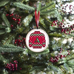 May include: A Christmas tree ornament featuring a pixelated red sweater design with a green letter "A". The ornament is framed in a wooden embroidery hoop and hangs from a red and black plaid ribbon. The background shows a Christmas tree with red berries.