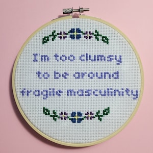 May include: A cross-stitch embroidery hoop with the text "I'm too clumsy to be around fragile masculinity" in purple thread. The design includes floral accents in green, blue, and purple. The hoop is made of wood and set against a pink background.