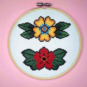 May include: A white embroidery hoop with a wooden frame, featuring two embroidered flowers. The top flower is yellow with a blue centre and green leaves. The bottom flower is red with a black centre and green leaves.