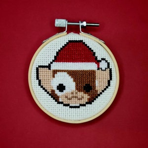 May include: A white and brown cross-stitch embroidery of a cartoon elf wearing a red Santa hat. The embroidery is framed in a wooden hoop.