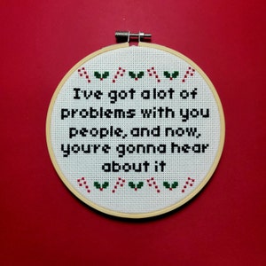 Puede incluir: Un aro de bordado blanco con fondo rojo. El aro tiene una tela blanca con texto negro que dice "Tengo muchos problemas con ustedes, la gente, y ahora, van a escuchar sobre esto". El texto está rodeado de diseños de bastones de caramelo rojos y verdes y acebo.