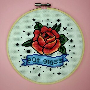 Puede incluir: Arte de bordado en aro con una rosa roja con hojas verdes y una pancarta azul con el texto "But Gross". El fondo es blanco con estrellas negras.