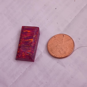May include: A rectangular, polished gemstone with a vibrant mix of red, orange, and purple hues. The gemstone is next to a U.S. penny for size comparison. The penny is dated 2022.