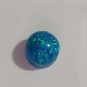 May include: A round, blue opal gemstone with flecks of green and blue throughout. The gemstone is smooth and polished, with a glossy finish. The opal is set against a white background.