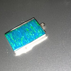 May include: Rectangular pendant with a vibrant blue and green opal-like stone set in a silver-colored frame. The pendant has a small loop at the top for a chain. The stone has a shimmering, iridescent quality.