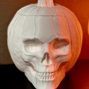 May include: White ceramic pumpkin shaped like a skull with a detailed face. The pumpkin is lit from the side, casting a warm orange glow on one side.