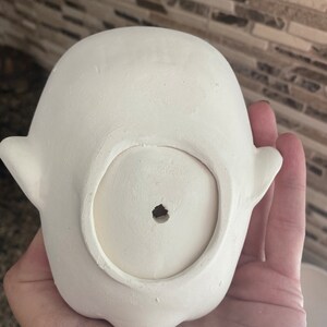 May include: A small, unpainted ceramic ghost-shaped planter. The planter has a round opening at the bottom and small, wing-like protrusions on either side. The surface is smooth and ready for painting.