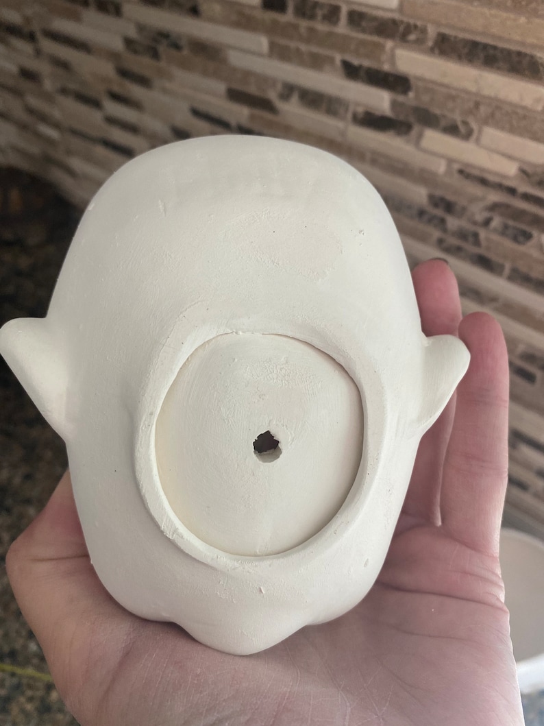 May include: A small, unpainted ceramic ghost-shaped planter. The planter has a round opening at the bottom and small, wing-like protrusions on either side. The surface is smooth and ready for painting.