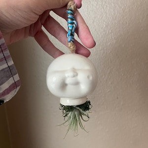 May include: A white ceramic face planter with a smiling expression, suspended by a natural rope with blue and silver accents. The planter holds a small air plant and moss, creating a unique hanging decor piece.