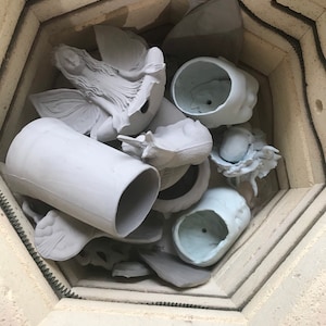 May include: A selection of unpainted ceramic sculptures and pottery items inside a kiln. The collection includes a fairy figurine, face-shaped planters, and other decorative items. The pieces are in various stages of completion, ready for firing and glazing.