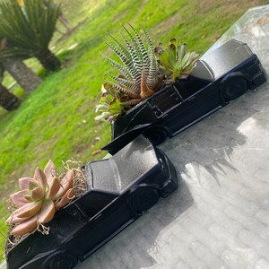 May include: Two black car-shaped planters with succulents growing inside. The planters are made of a smooth, matte black material and have a realistic car design. The succulents are a variety of green and brown colors.