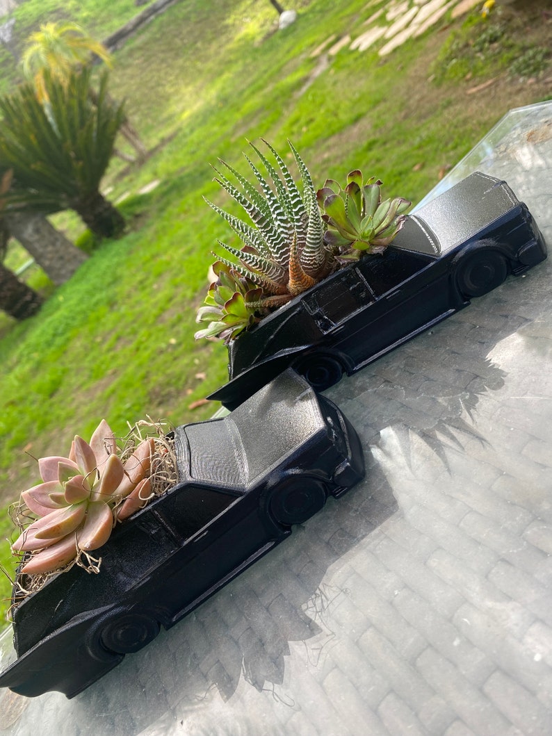May include: Two black car-shaped planters with succulents growing inside. The planters are made of a smooth, matte black material and have a realistic car design. The succulents are a variety of green and brown colors.
