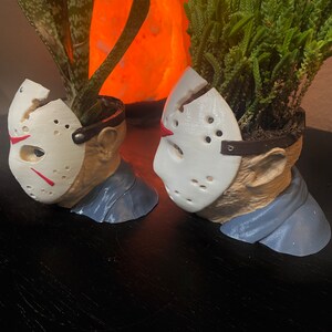Puede incluir: Dos macetas de cer&aacute;mica blanca con forma de la cabeza de Jason Voorhees de la franquicia de pel&iacute;culas de terror Viernes 13. Cada maceta tiene una peque&ntilde;a planta verde que crece en la parte superior.