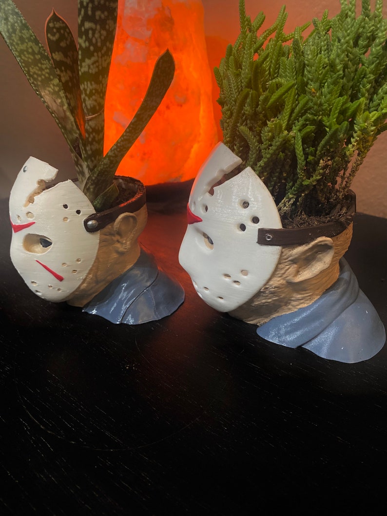 Puede incluir: Dos macetas de cer&aacute;mica blanca con forma de la cabeza de Jason Voorhees de la franquicia de pel&iacute;culas de terror Viernes 13. Cada maceta tiene una peque&ntilde;a planta verde que crece en la parte superior.