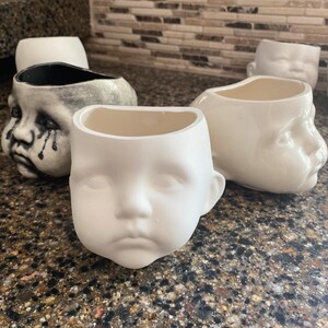 May include: A collection of ceramic planters, each moulded in the shape of a baby's face. One is painted grey with tear streaks, the others are plain white. The planters are designed to hold plants or other items. They sit on a speckled countertop.