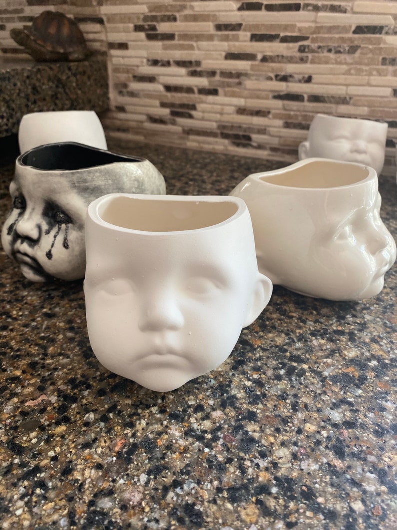 May include: A collection of ceramic planters, each moulded in the shape of a baby's face. One is painted grey with tear streaks, the others are plain white. The planters are designed to hold plants or other items. They sit on a speckled countertop.