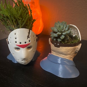 Può includere: Due vasi da fiori stampati in 3D a forma di teste di personaggi di film horror. Il vaso a sinistra è bianco e a forma di maschera da hockey di Jason Voorhees. Il vaso a destra è beige e a forma di testa umana con una sciarpa blu. Entrambi i vasi hanno piccole piante che crescono al loro interno.