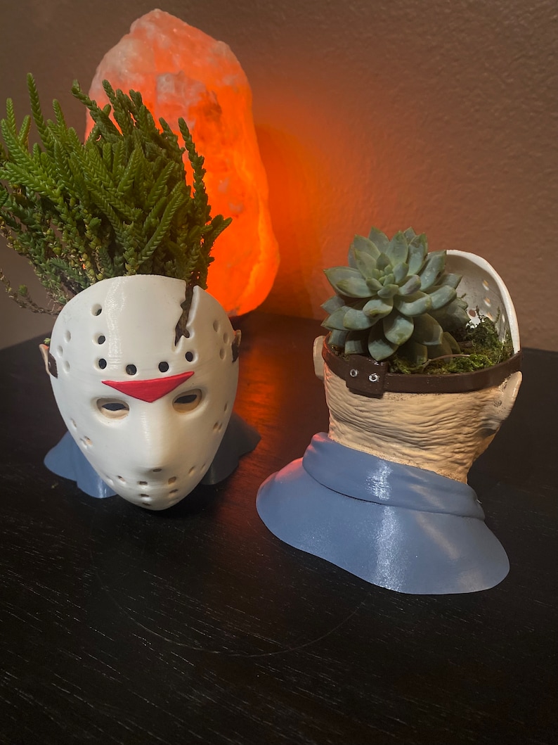 Puede incluir: Dos macetas impresas en 3D con forma de cabezas de personajes de pel&iacute;culas de terror. La maceta de la izquierda es blanca y tiene forma de la m&aacute;scara de hockey de Jason Voorhees. La maceta de la derecha es beige y tiene forma de cabeza humana con una bufanda azul. Ambas macetas tienen peque&ntilde;as plantas que crecen en ellas.
