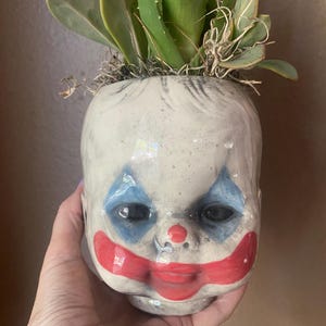 May include: A ceramic planter shaped like a doll head with a glossy off-white finish. The face features painted blue diamond shapes around the eyes, a red nose, and a wide red mouth. Green succulent plants are growing out of the top of the head.