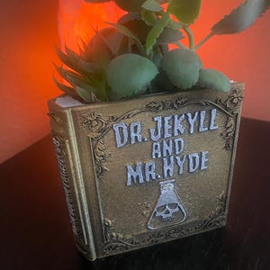 以下が含まれることがあります： 「ジキルとハイド」と題された金のブック型のプランター。プランターには緑色の多肉植物が植えられています。