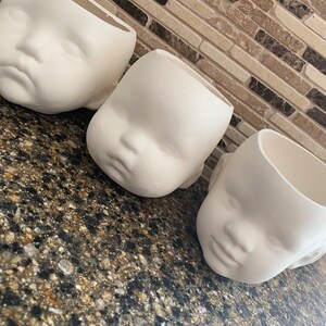 May include: Three off-white ceramic planters shaped like baby faces. The planters have a matte finish and are arranged on a speckled countertop. The planters are open at the top, ready for planting.