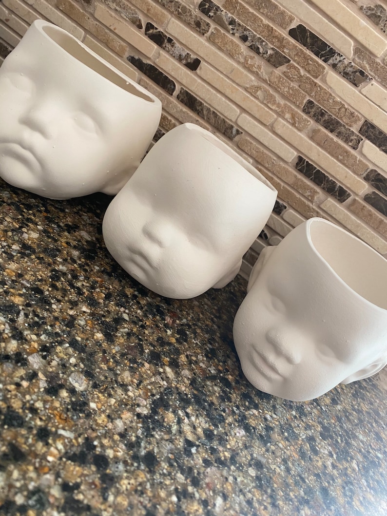 May include: Three off-white ceramic planters shaped like baby faces. The planters have a matte finish and are arranged on a speckled countertop. The planters are open at the top, ready for planting.