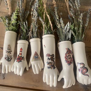 Pode incluir: Seis vasos de parede de cerâmica em tom off-white com formato de mão, cada um com um gráfico colorido estilo tatuagem, incluindo pin-up, farol, olho da providência, dragão, caveira e cobra. Cada vaso contém ramos de lavanda e alecrim secos.