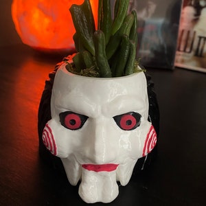 May include: A white ceramic planter shaped like the face of the character Jigsaw from the Saw horror movie franchise. The planter has black hair, red eyes, and a red spiral pattern on the cheek. A small green succulent plant is growing in the planter.