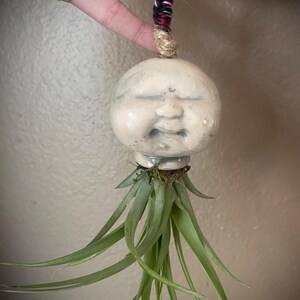 May include: A ceramic head sculpture with a smooth, off-white finish and a serene facial expression, featuring closed eyes and a small mouth. A green air plant is attached to the base, and a braided cord in black, white, and red hangs from the top.