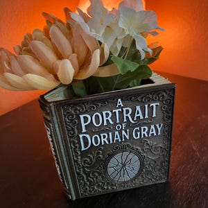 May include: A decorative book-shaped vase with the title "A Portrait of Dorian Gray" in white lettering. The vase is filled with peach and white artificial flowers. The vase has an ornate, vintage design and is made of a dark brown material.