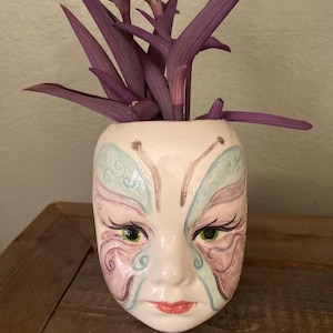 Puede incluir: Una maceta de cerámica blanca con forma de rostro, con ojos verdes, labios rojos y detalles decorativos azules y rosas. La maceta contiene una planta morada con hojas largas y delgadas. La maceta está sobre una superficie de madera.