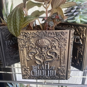 May include: A gold-colored book planter with a detailed design and the words "The Call of Cthulhu" on the front. The planter is filled with green plants.