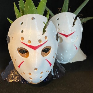 May include: Two white, 3D-printed masks with red details and eye holes, resembling a hockey mask, each holding a green aloe vera plant. The masks sit on a dark blue base against a black background. The plants have green, spiky leaves.
